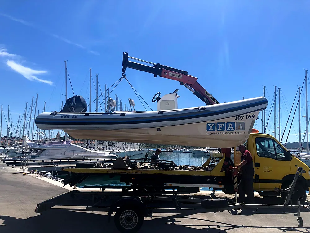 Inflatable boat in the crane slings above a trailer in the marina